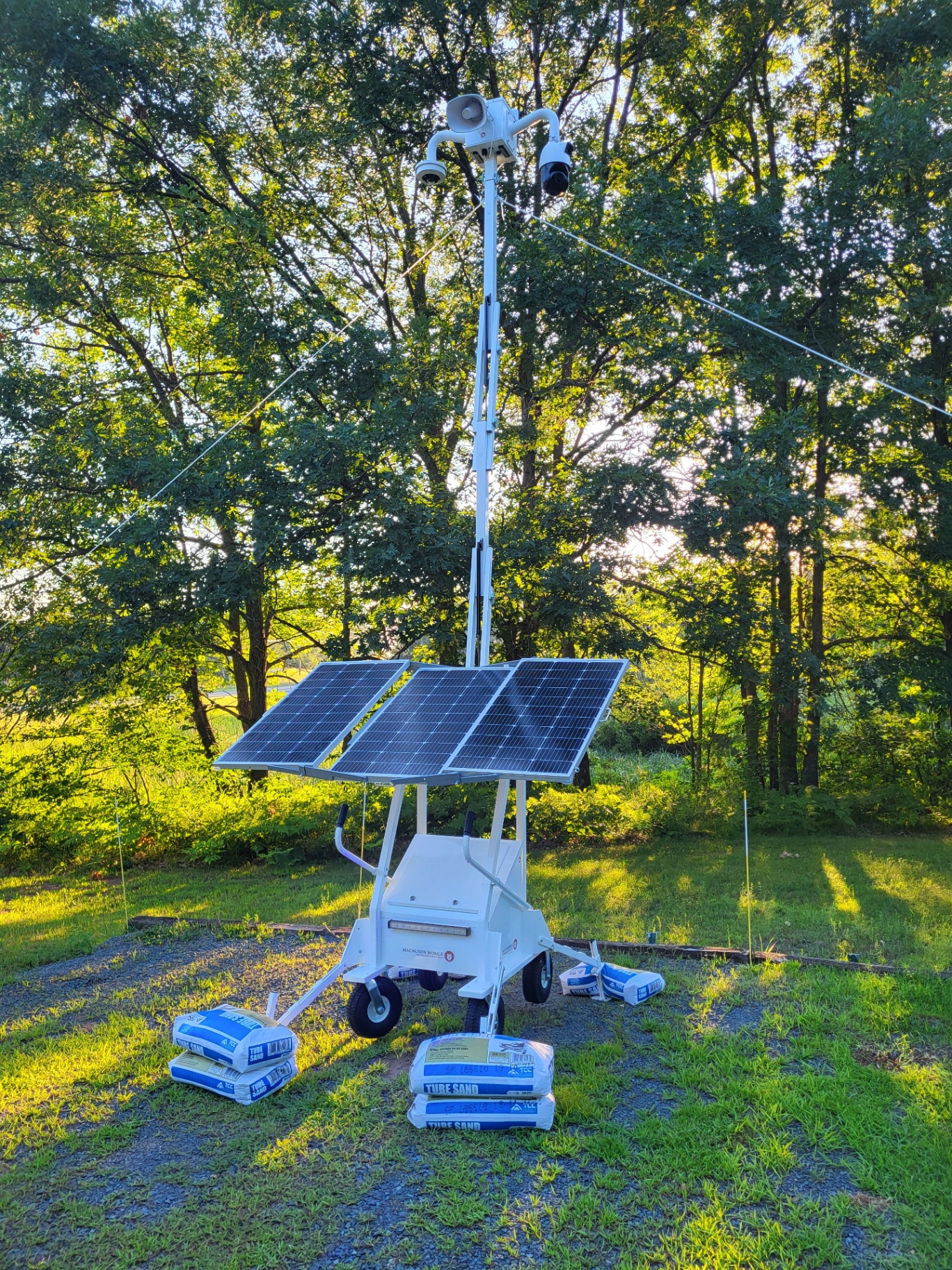 Magnuson mobile solar surveillance trailer deployed in a wooded field with mast raised, PTZ cameras, speaker, and three solar panels, secured with tube sand ballast.